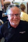 Older man with glasses and sunglasses on head smiling in a busy restaurant setting wearing a black polo shirt.