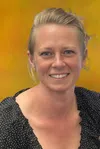 Smiling woman with blonde hair wearing a black polka dot blouse against a warm yellow background