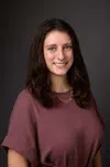 Smiling young woman with long dark hair wearing a mauve top and necklace against a dark background