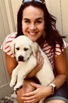 Smiling woman in striped shirt holding a white Labrador puppy outdoors near a fence.