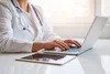 Doctor wearing white coat typing on laptop with tablet on white desk under bright window light.