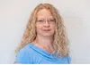 Portrait of a woman with curly blonde hair wearing glasses and a blue top against a plain light background