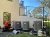 Estes Services Technician in orange shirt installing three Carrier outdoor air conditioning units beside a modern house on sunny day