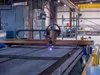 Industrial CNC plasma cutting machine cutting metal sheets in a factory workshop with safety barriers.