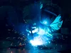 A welder in protective gear works on metal, creating bright sparks against a dark background.