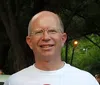 Smiling man with glasses wearing a white t-shirt standing outdoors near trees and street lamps at dusk
