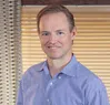 Smiling male dentist in blue checkered shirt standing indoors with patterned curtain and window blinds in background.