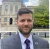 Man with a beard in suit and tie standing outdoors near a historic building and greenery on a sunny day