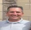 Smiling man in light gray polo shirt standing outside against a beige and brick wall background.