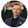 Man in blue blazer and black shirt standing in front of a blurred company logo background.