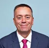 Smiling man in blue suit and pink tie against a light blue background, professional headshot portrait