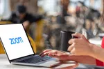 Person holding black mug and typing on laptop with Zoom logo on screen in a bright cafe setting
