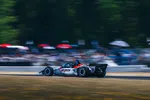 IndyCar race car number 15 speeding on track with blurred crowd and trees in background