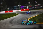 IndyCar race cars speed around a curved track with Firestone banners and spectators in the background.