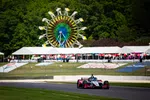 IndyCar race car speeding on track with colorful Ferris wheel and tents in background during event