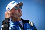 Race car driver in blue racing suit adjusts earpiece under clear blue sky, wearing white cap and sunglasses