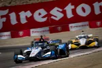 IndyCar race cars speeding on track with Firestone sponsor banners in the background at a racing event.