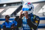 Race car driver adjusts helmet before a racing event with team members in blue uniforms in the background.