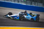Blue and white IndyCar racing on an asphalt track with advertising logos and a blue wall background