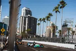 IndyCar race cars speeding on an urban circuit track lined with palm trees and skyscrapers under clear blue sky