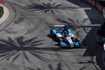 IndyCar racing car navigating a sharp turn on track with large palm tree shadows overhead in bright daylight