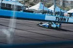 IndyCar racing car speeds past Turn 1 on a sunny race track with tent structures in the background