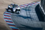 IndyCar race car navigating a sharp turn on a racetrack with red, white, and blue curbs under bright sunlight.