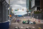 Open-wheel race cars competing on a city street circuit with barriers, fences, and traffic lights overhead.
