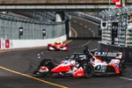 Red and white IndyCar racing on a curving track with barriers, closely followed by another race car.