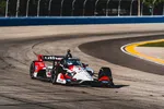 Red, white, and black IndyCar racing on curved asphalt track with blue fencing in background.