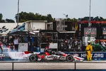 Race car speeding on track with crew and spectators behind safety fence at motorsports event.