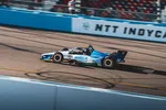IndyCar race car sponsored by WWEX Group speeding on the track with NTT IndyCar banner in background.
