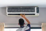 Technician inspecting and repairing a wall-mounted air conditioner unit indoors using a handheld tool.