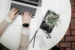 Woman typing on a laptop with green plant, phone, glasses, and stationery on a table.