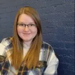 Smiling young woman with long red hair and glasses wearing a plaid shirt against dark blue brick wall