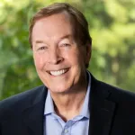 Smiling middle-aged man wearing a blue shirt and dark blazer with a blurred green outdoor background