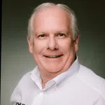 Smiling elderly man with white hair wearing a white collared shirt against a gray background