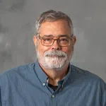 Portrait of an older man with gray hair and beard wearing glasses and a blue checkered shirt against a gray background