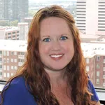 Smiling woman with long red hair and blue eyes wearing a blue top in an urban office setting with buildings behind.