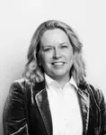 Smiling woman in velvet blazer and white shirt standing against plain background in black and white photo.