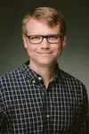 Smiling man with glasses wearing a black and white checkered shirt against a plain background.