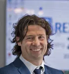 Smiling man with curly hair wearing a blue suit and white shirt in an office setting.