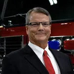 Middle-aged man in suit and red tie smiling, standing in front of a fire truck with emergency lights.