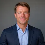 Professional headshot of a confident man in a blue suit jacket and light blue shirt against a gray background