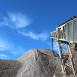Conveyor belt dumping gravel onto large aggregate pile under bright blue sky with scattered clouds.
