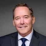 Professional headshot of a middle-aged man in a navy suit, white shirt, and blue tie against gray background