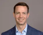 Smiling man with short brown hair wearing blue blazer and light blue shirt against gray background