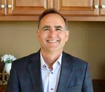 Smiling middle-aged man in a blue plaid blazer and white shirt standing in front of wooden cabinets.