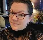 Woman with short hair and red glasses smiling indoors near office equipment and floral arrangement on a cabinet.