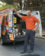 Estes Services Technician in orange uniform standing next to open service van with tools and equipment inside on sunny day.